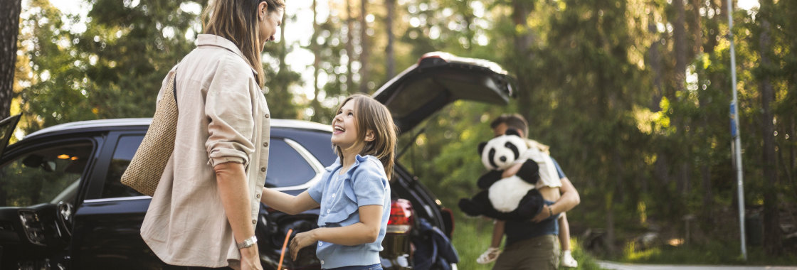 Mother and daughter standing beside an electric vehicle being charged whilst father unloads second child and large panda soft toy from vehicle